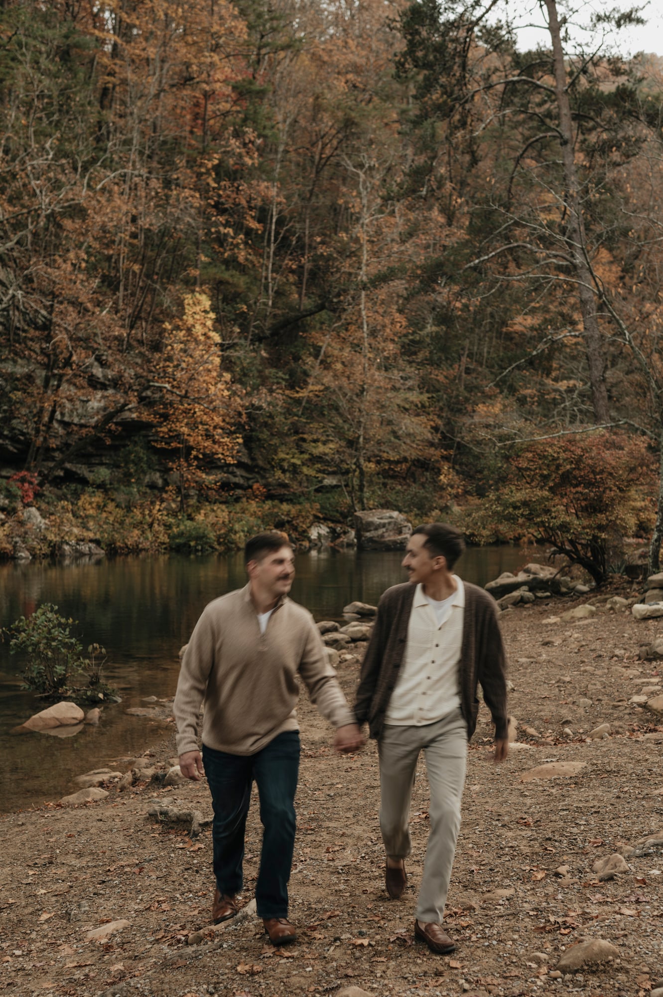 Engaged couple walking hand in hand along a rocky riverbank in autumn