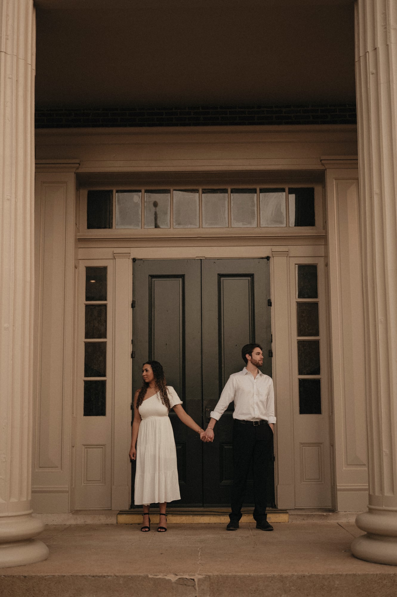 Engaged couple holding hands in front of tall wooden doors flanked by columns