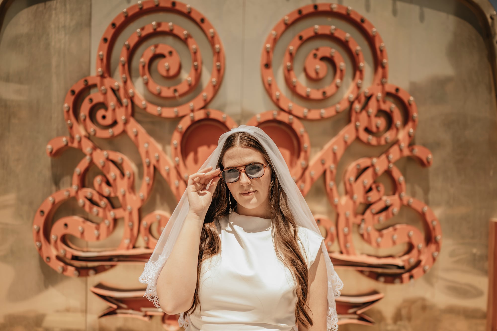 Bride in a short white dress and veil adjusting sunglasses in front of a vintage sign