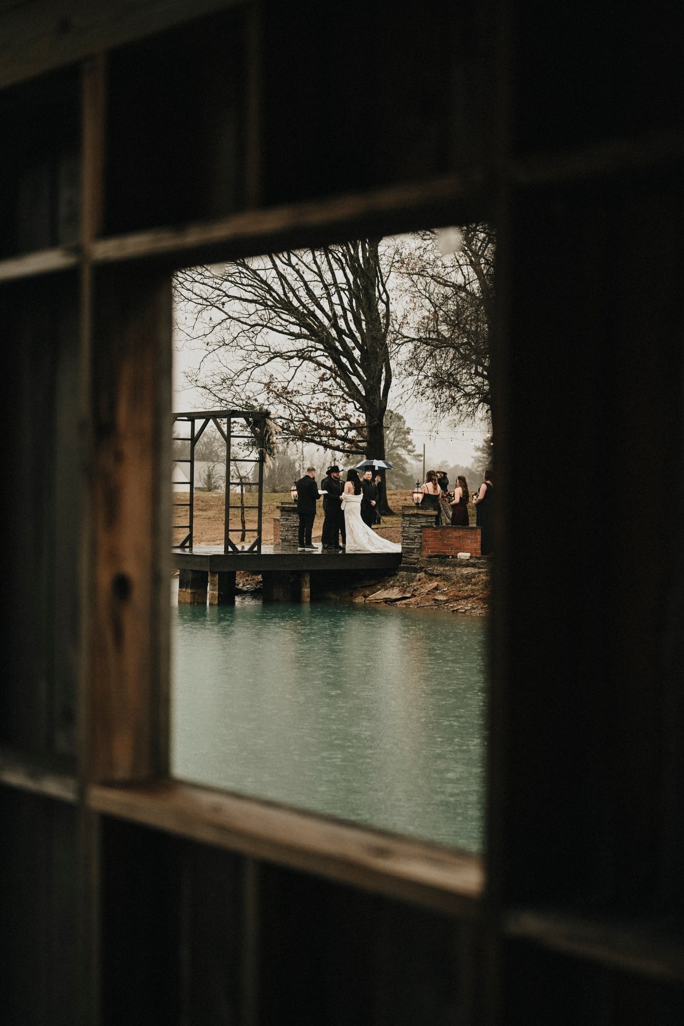 Wedding ceremony on a lakeside dock viewed through a barn window on a rainy day