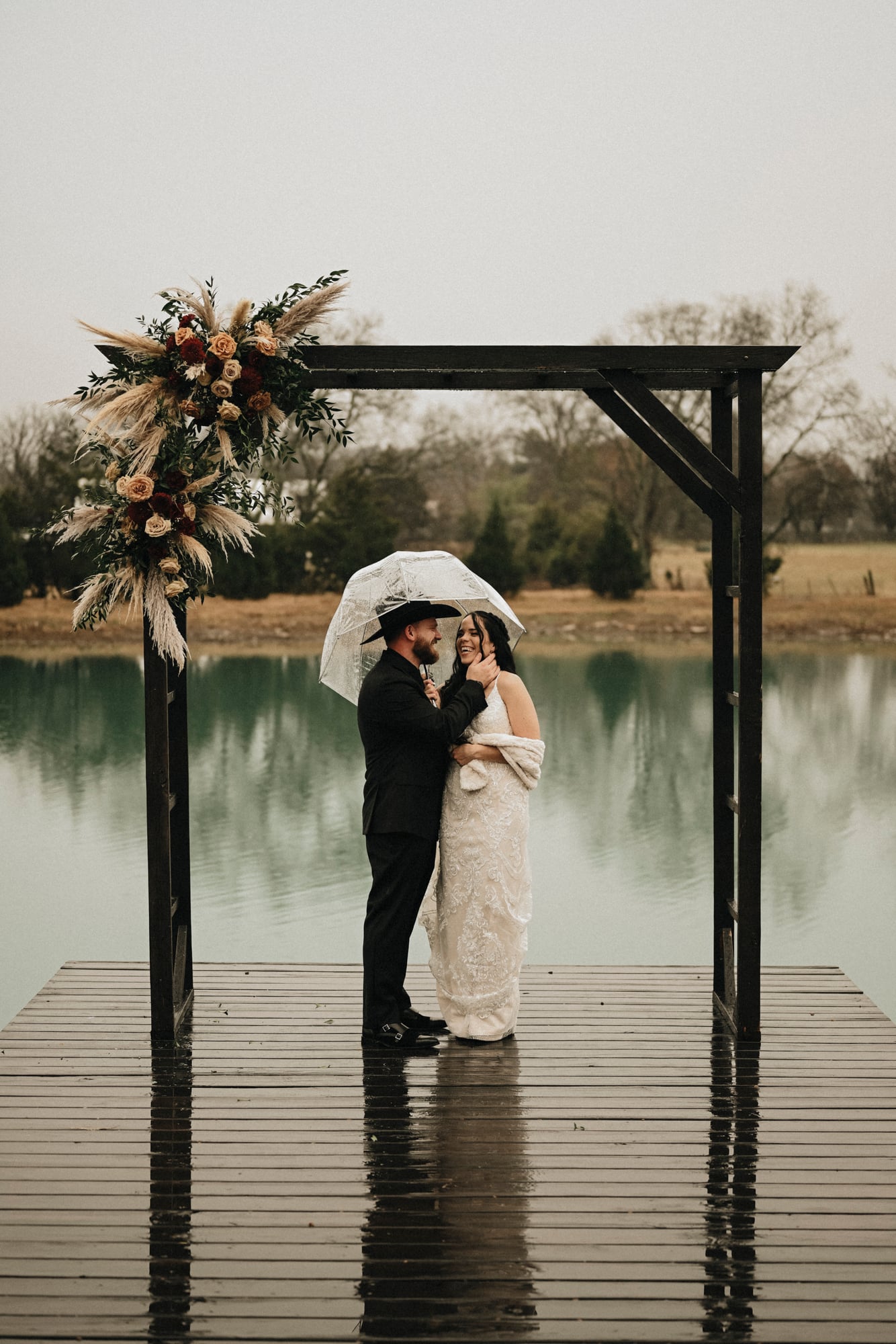 Bride and groom standing under a floral arch with a clear umbrella on a rainy dock